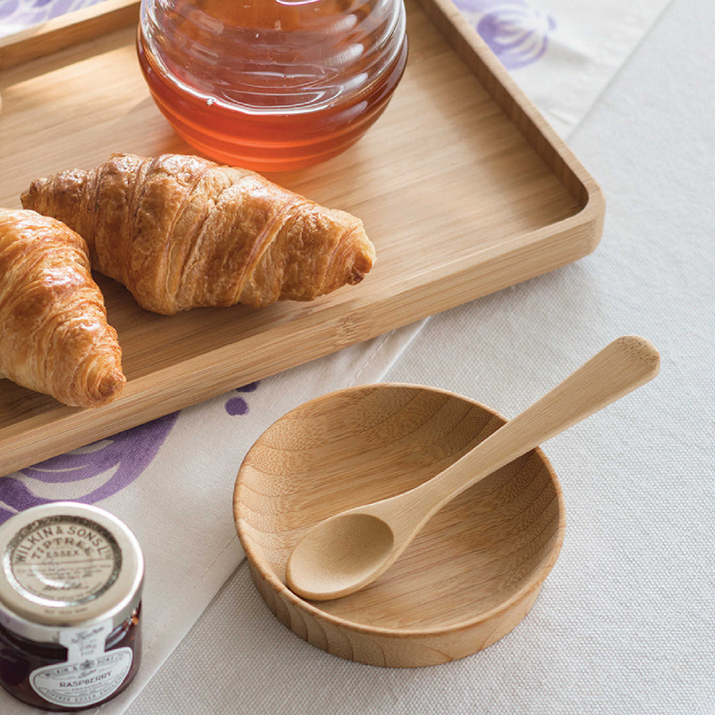 A large bamboo condiment cup is shown with a bamboo teaspoon and bamboo tray. There is a jar of honey and a fresh croissant on the tray, and a small jar of jam is on the table.