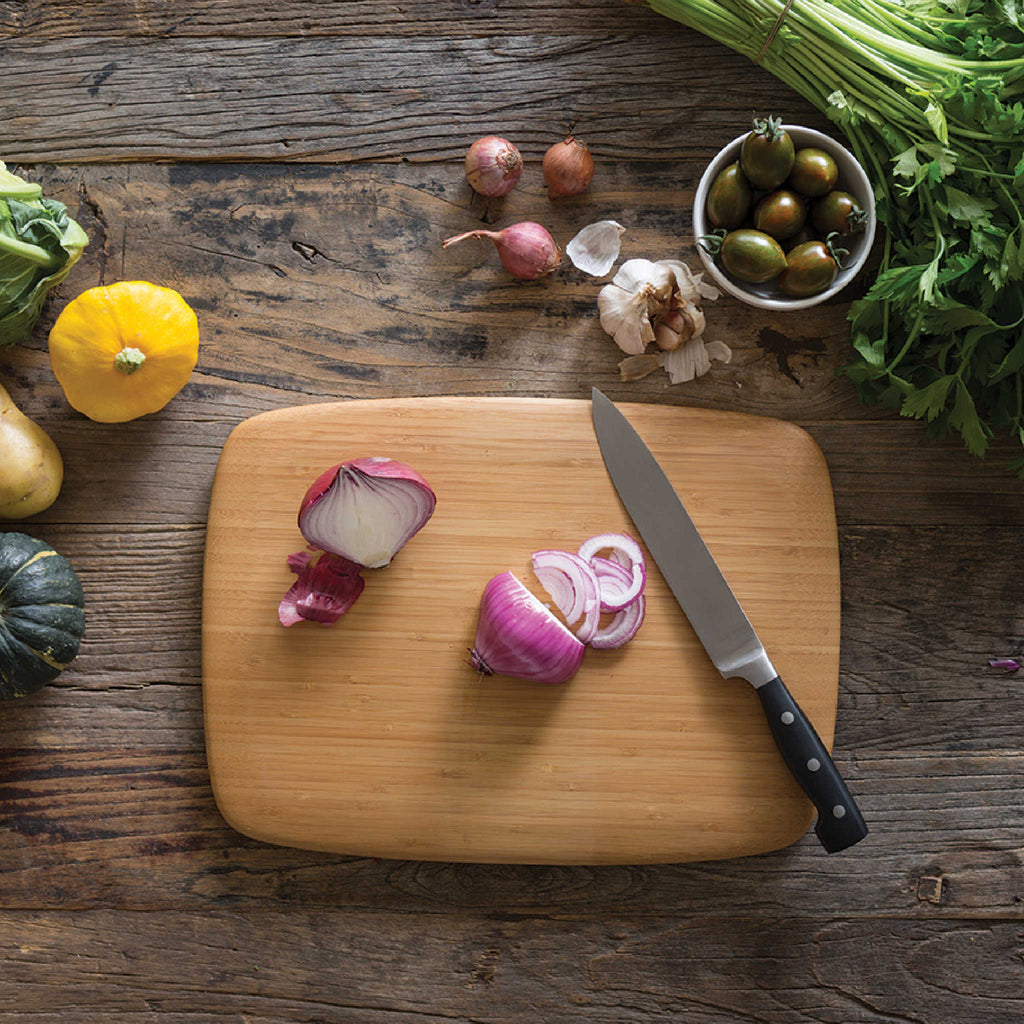 A medium Classic Cutting & Serving Board is used to slice an onion. There are more colorful veggies near the board waiting to be prepped.