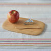 A bamboo bar board rests on a tablecloth. An apple, a wedge of cheese, and a bamboo spreader are atop the board.