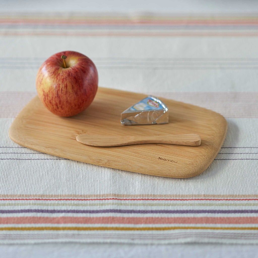 A bamboo bar board rests on a tablecloth. An apple, a wedge of cheese, and a bamboo spreader are atop the board.