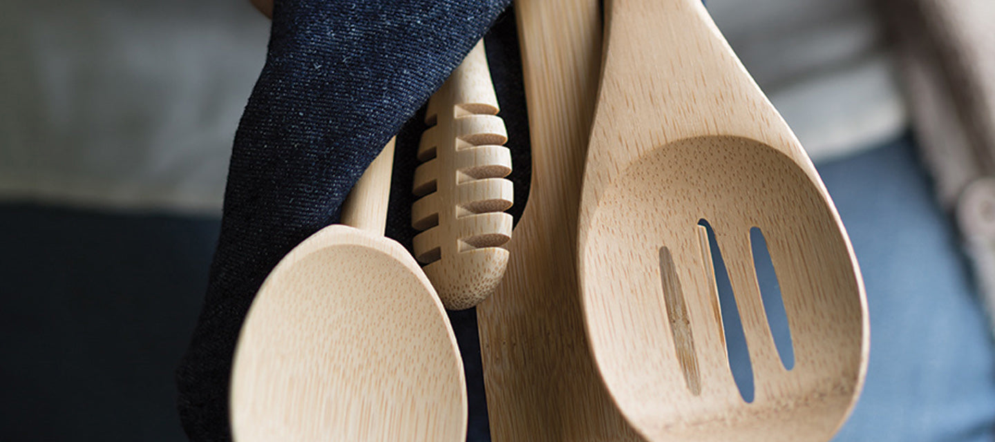 Bamboo utensils are held together with a dishtowel. Featured is a bamboo slotted spoon, a bamboo honey dipper, and a bamboo mixing spoon.