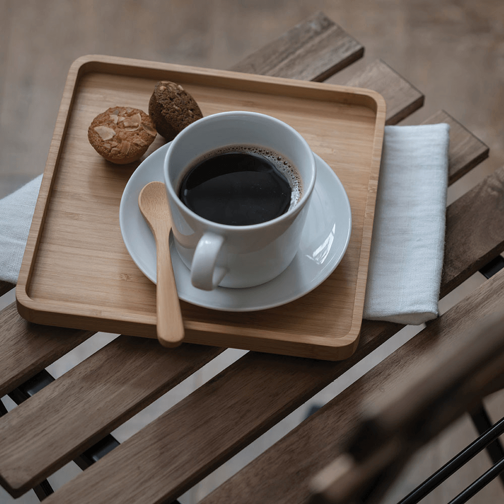 A Teaspoon is resting on the saucer of a coffee cup. The cup and saucer are on a bamboo serving tray.with tea on tray