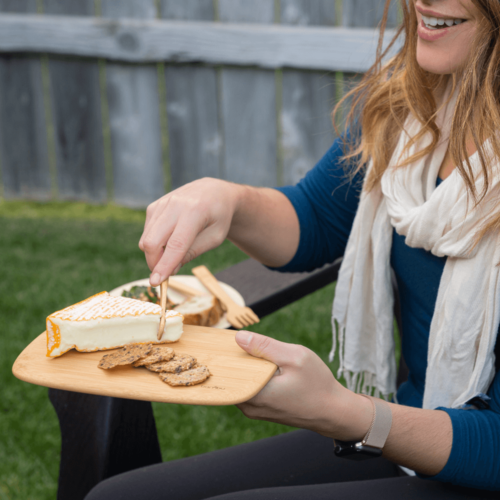 A woman with a white scarf and blue shirt uses a bamboo bar board to cut cheese to serve on crackers.