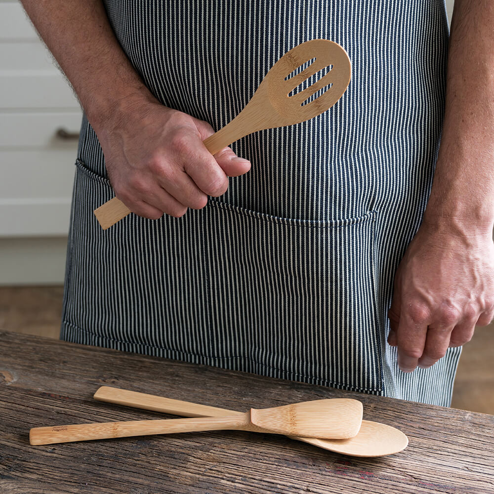 A person wearing a striped apron holds a Give it a Rest Slotted spoon. A Give it a Rest Spatula and Spoon are on the table in front of them.