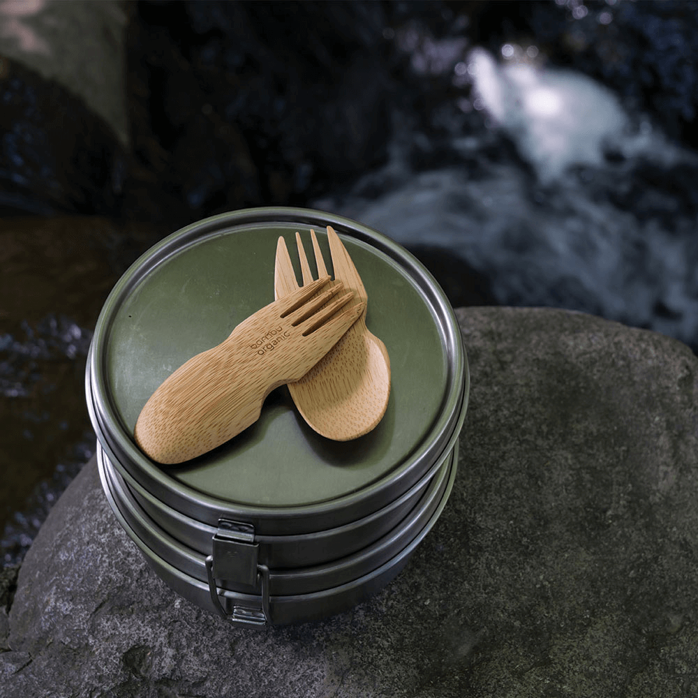 A pair of Bamboo Sporks are atop a lunchbox that is placed on a rock. There is a stream in the background.
