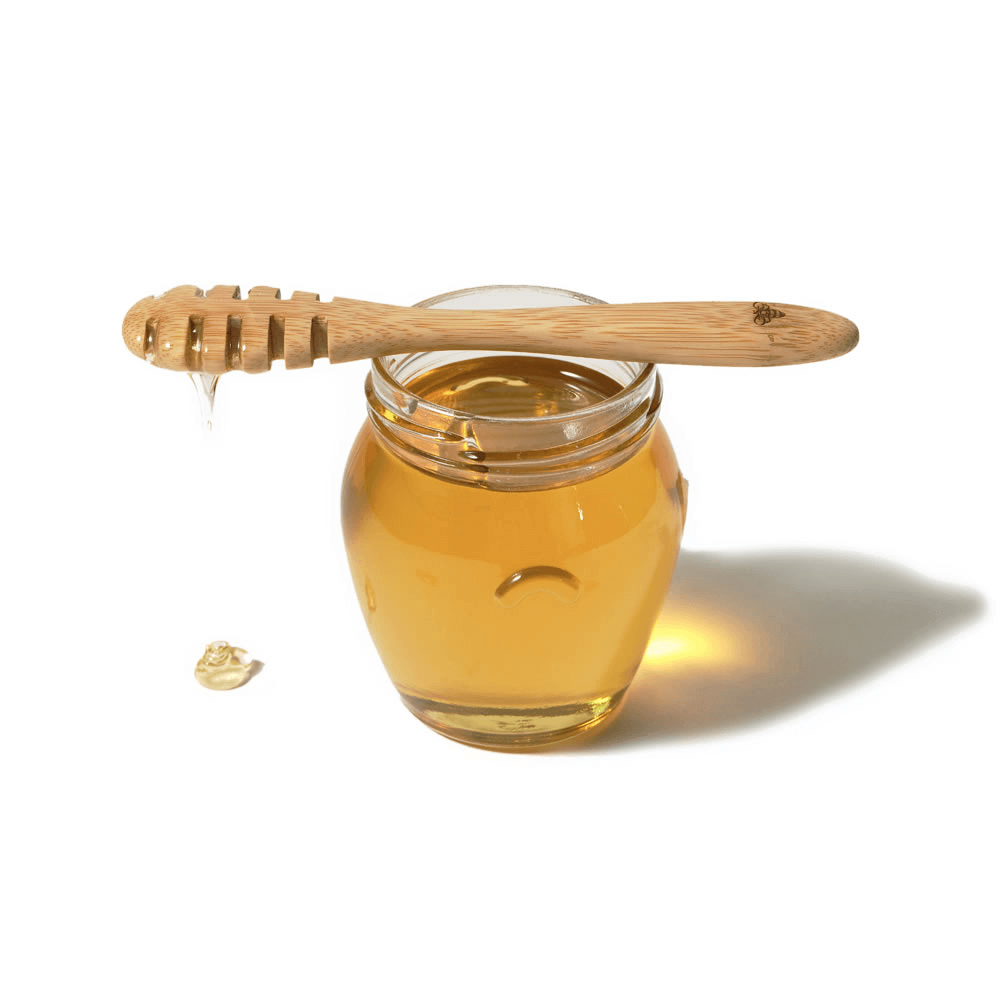 An Organic Bamboo Honey Dipper is laid across an open jar of honey. Honey drips from the utensil onto the countertop.