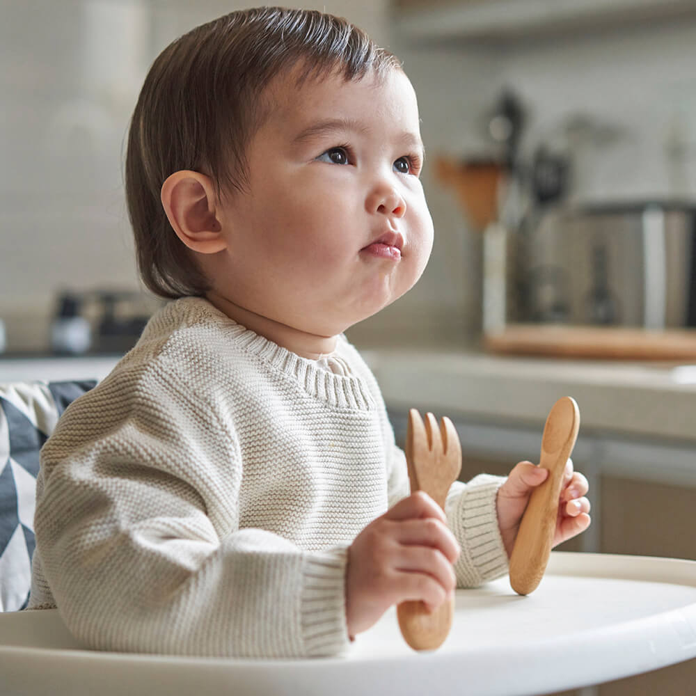 A young child sits in a high chair holding a Baby's/Toddler's Fork and Spoon Set (12M+) - bambu