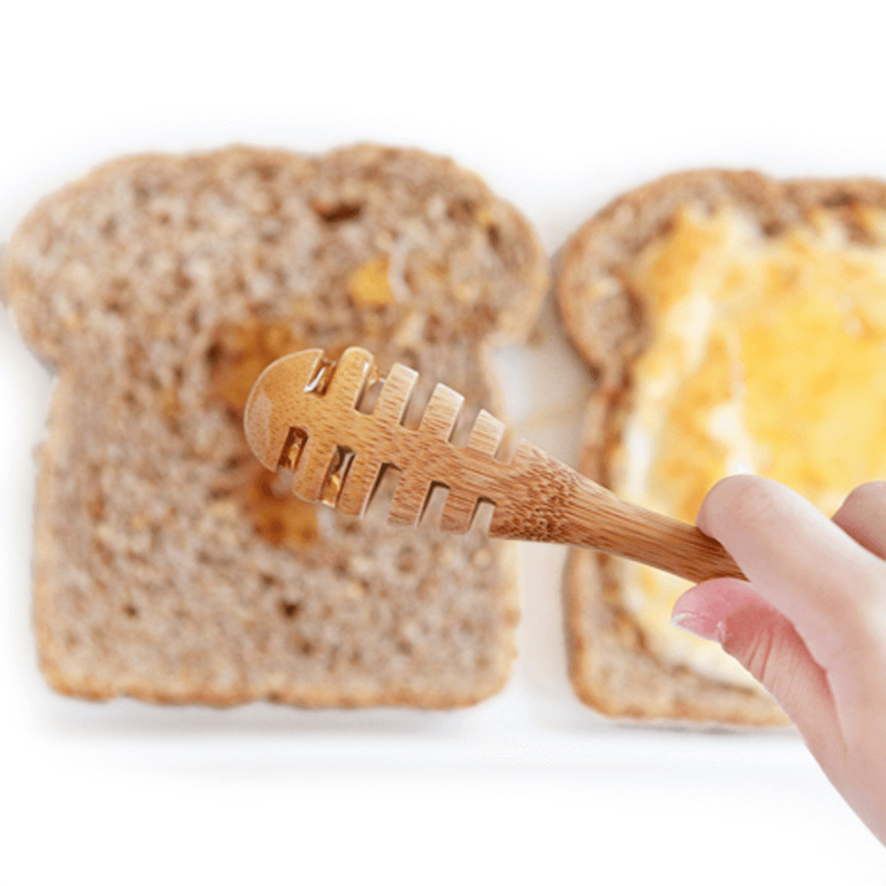 A hand holding an Organic Bamboo Honey Dipper drizzles honey onto bread. A second piece of bread awaits to complete the sandwich. 