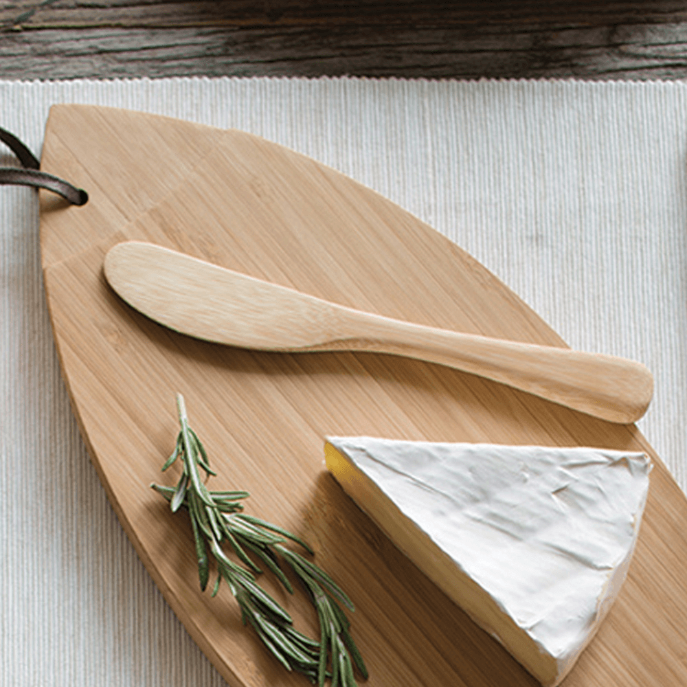 A 7" bamboo spreader is next to a wedge of brie, on a bamboo cutting board. There is a sprig of rosemary near the cheese.