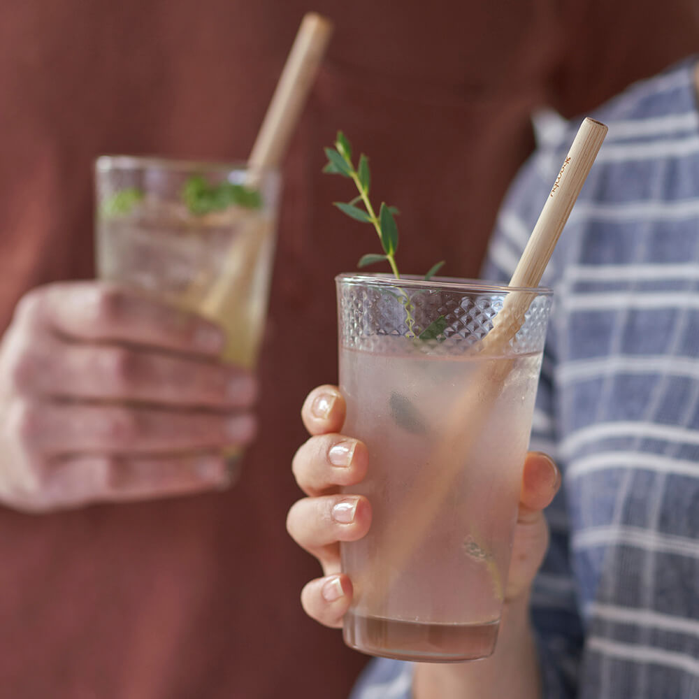 Two people hold drinking glasses, each glass has a Precision Reusable Bamboo Straw.