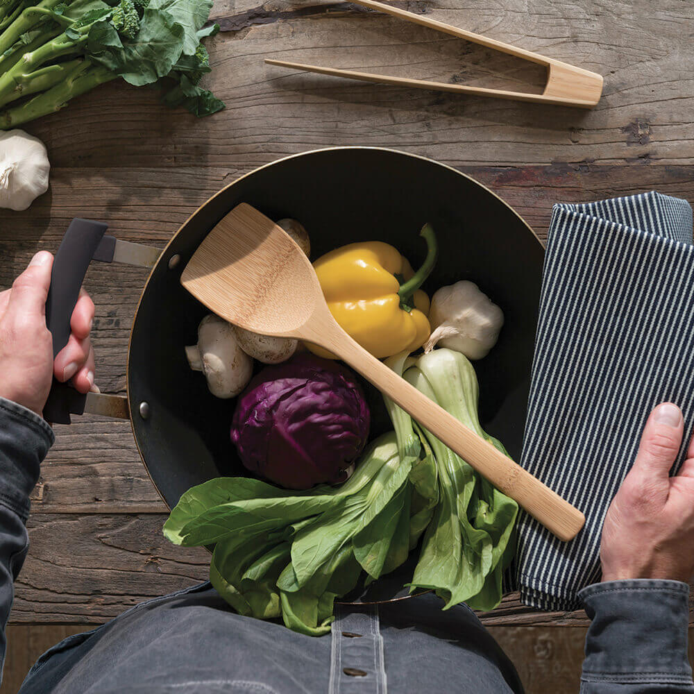 A Bamboo Wok Spatula is carried in a wok with colorful vegetables. A pair of tongs are on the counter next to more veggies.