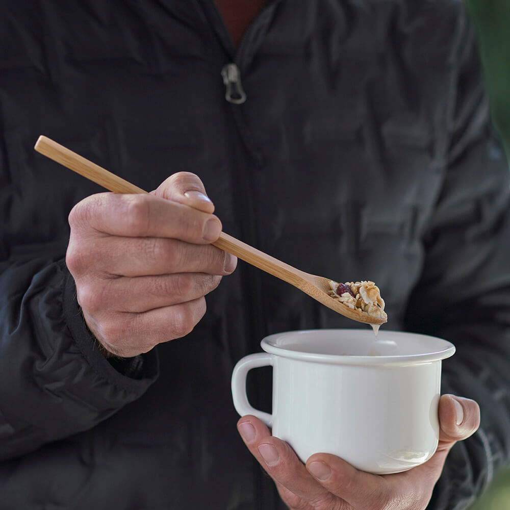 A person uses a bamboo trail spoon to eat granola from a white camping mug- bambu