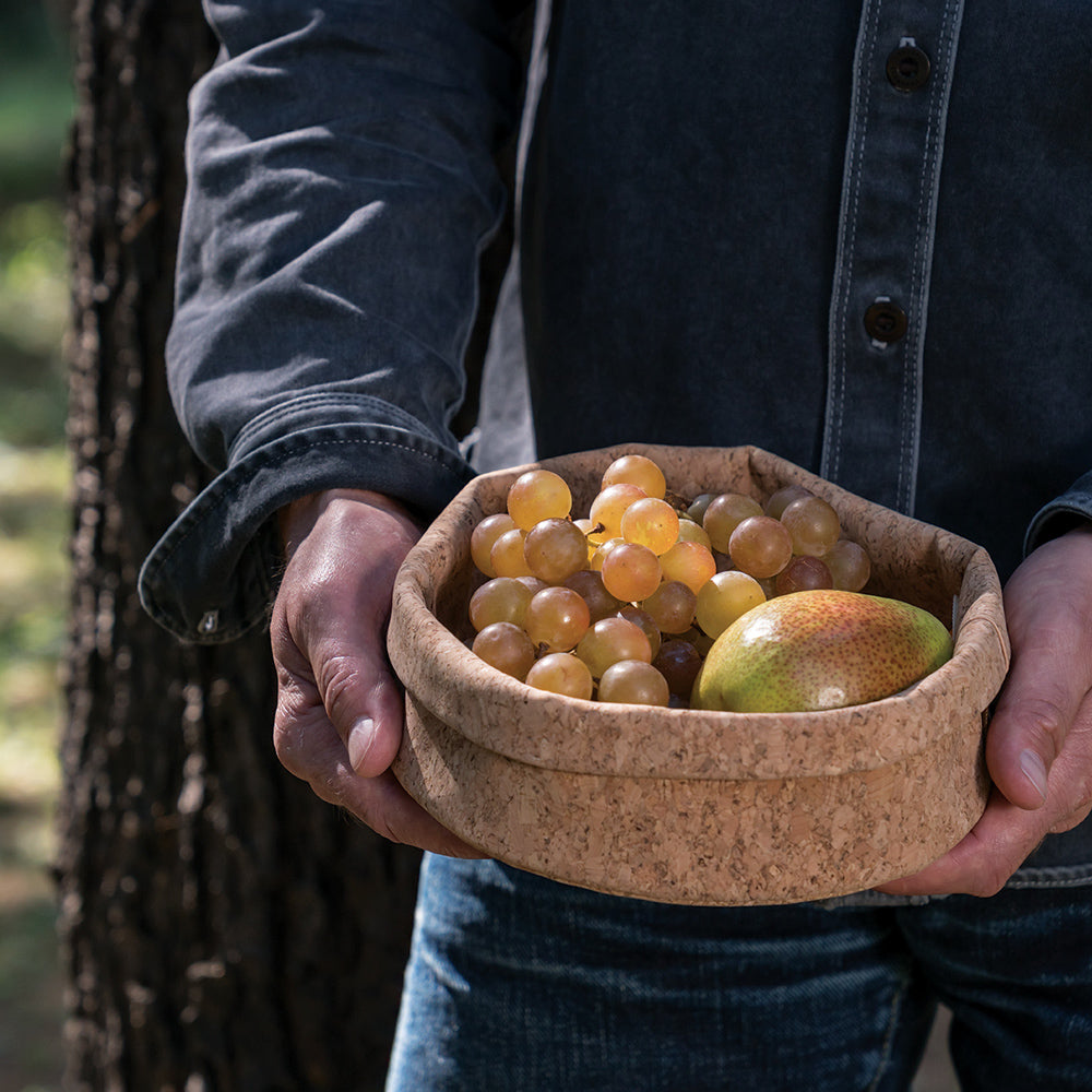 a person wearing a denim shirt is holding an 8-inch cork bowl. There are grapes and a pear in the bowl, and the edges of the bowl are folded down slightly.