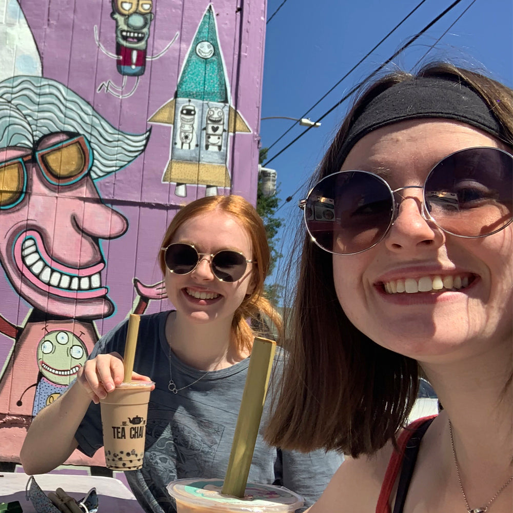 Two young women sit in front of a colorful mural. Each hold a Boba Tea with a Reusable Bamboo Jumbo Straw.