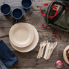 A stack of Veneerware® Round Bamboo Plates are next to Veneerware Cutlery and blue enamel camping mugs are on top of a wooden picnic table.