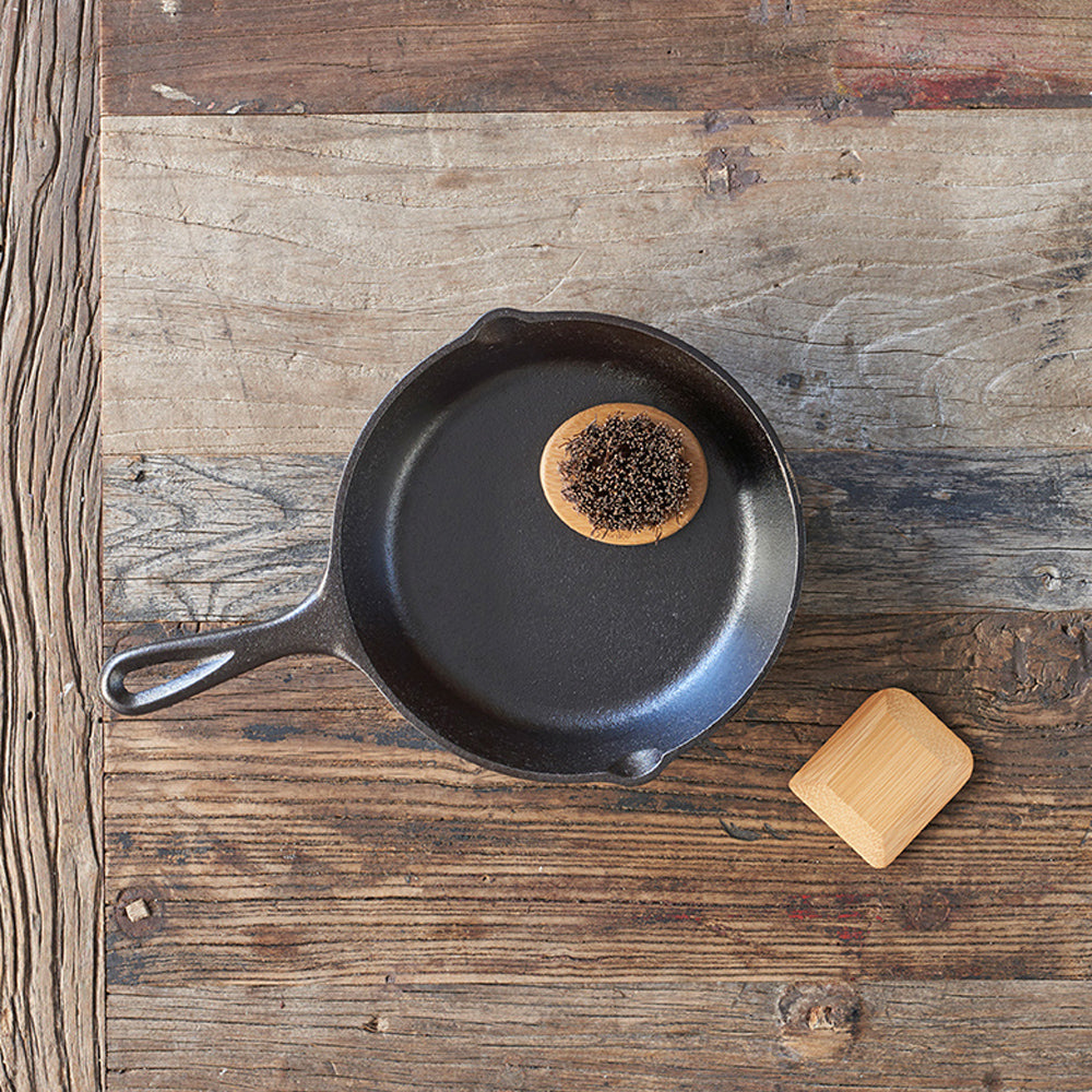 A Pot & Pan Scrub Brush sits in a cast iron pan. A pot scraper lays next to the pan on a wooden tabletop.