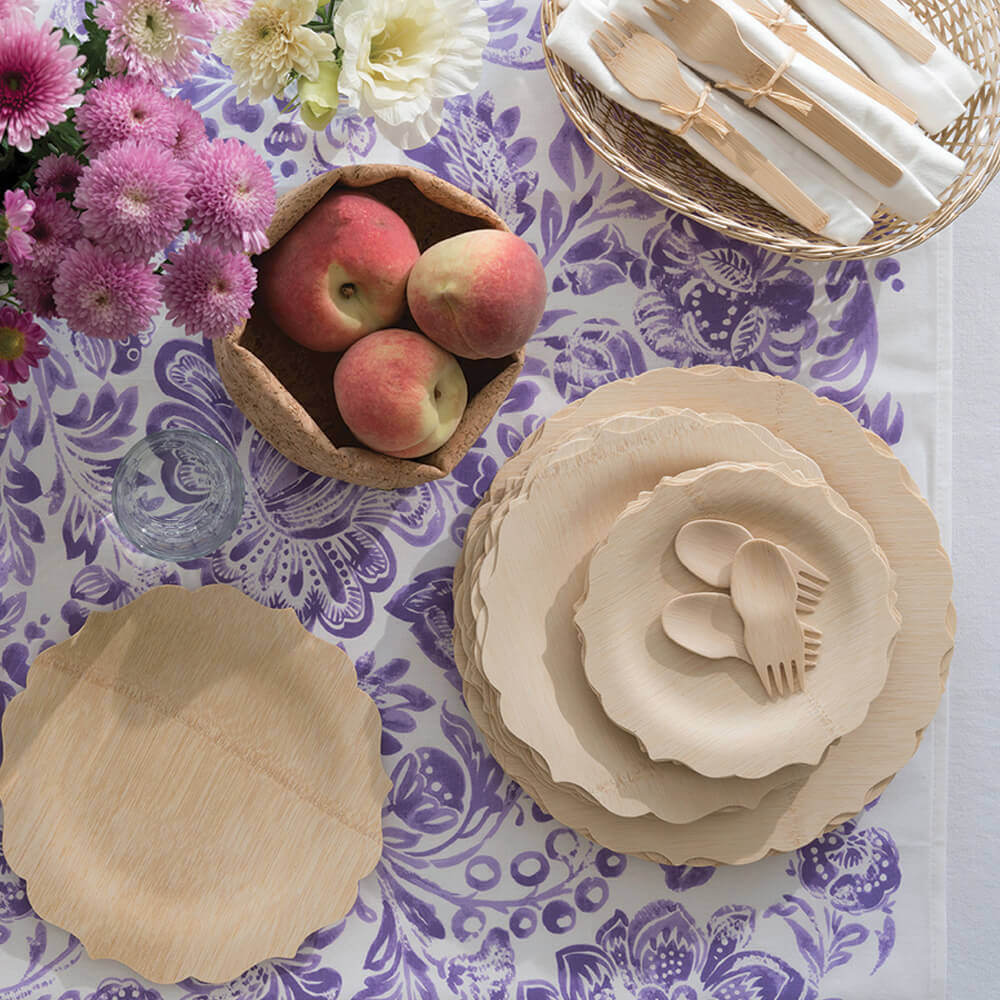 A stack of Veneerware® Fancy Bamboo Plates are atop a white and purple tablecloth. There is a basket of Veneerware cutlery nearby.