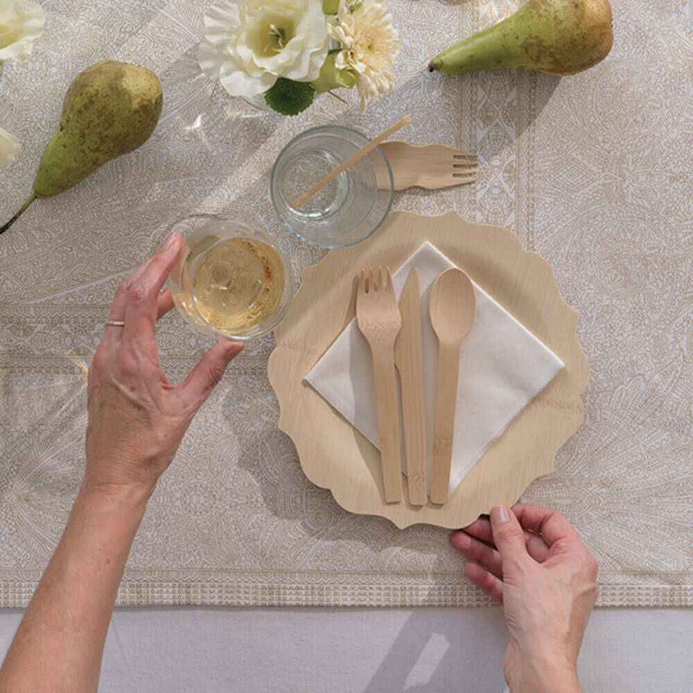 A 9" Fancy Veneerware plate is set on a table. There is a bamboo cocktail napkin and a set of cutlery on top of the plate. A person reaches into frame to pick up a wine glass set above the table.