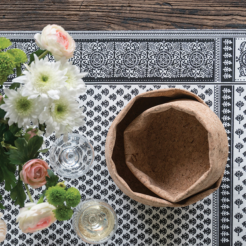 An 8 inch cork bowl is nested inside a 10 inch cork bowl. They are placed on a wood table that has a patterned tablecloth, and a vase of flowers is to the left.