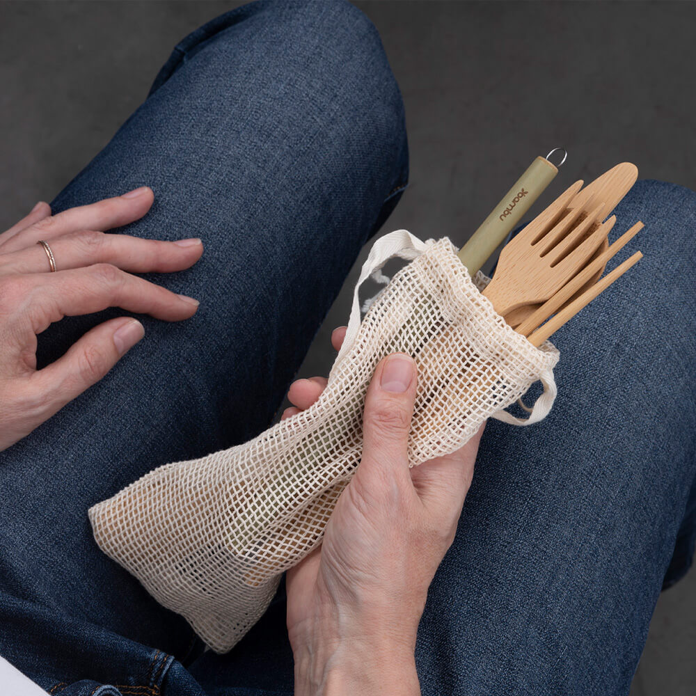 A person in blue jeans sits with an eat/drink tool kit in their hand. The drawstring bag is open so they utensils are peeking over the edge of the bag.