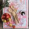 An All Purpose Bamboo Mixing Spoon is shown with other bamboo utensils on a pink floral table runner. A bowl of fruit is in the corner.
