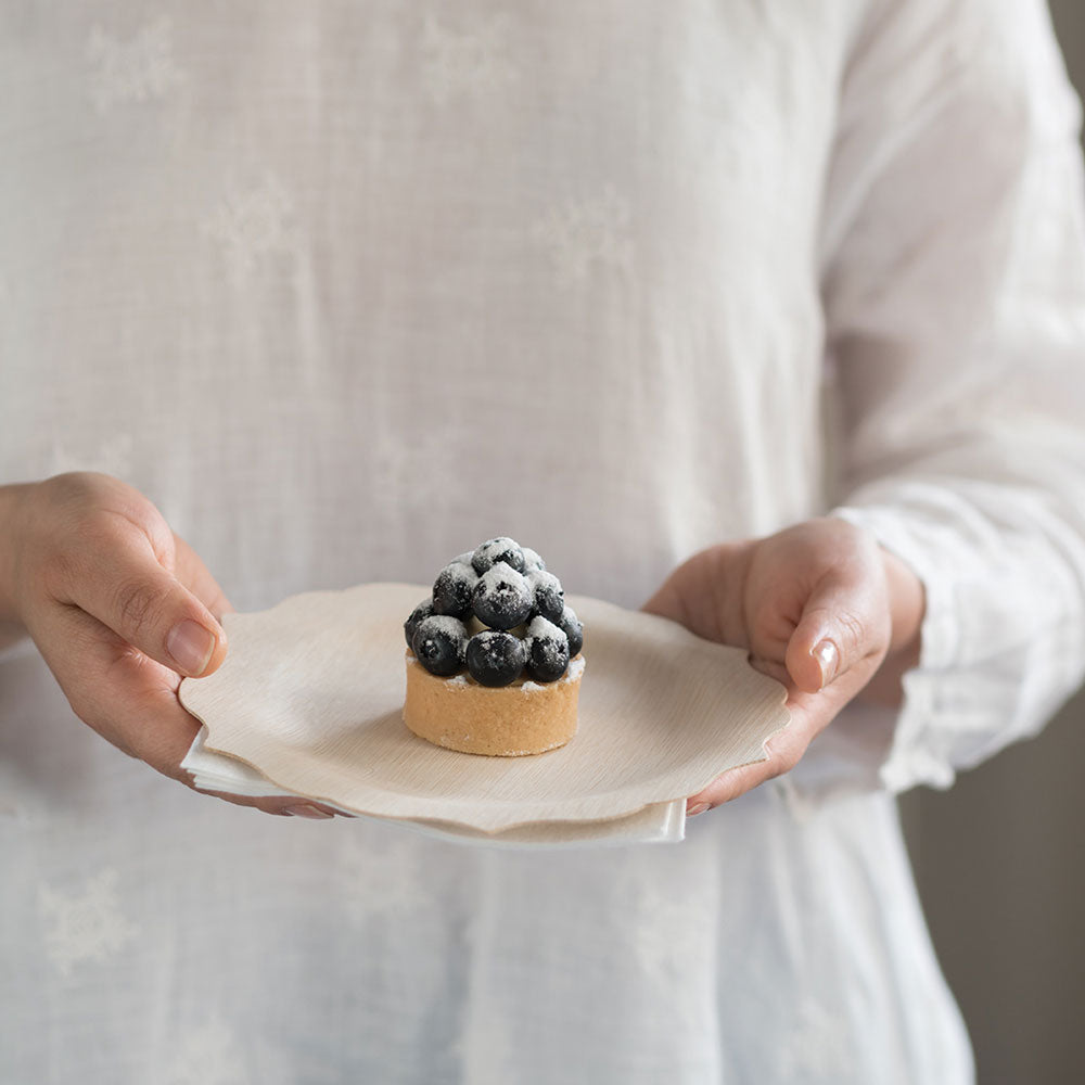 A person in a white blouse holds a 7" Veneerware® Fancy Bamboo Plate. A fruit tart is on the plate.