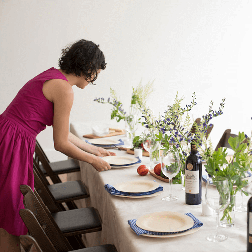 A woman in a pink dress sets a table for a holiday dinner. Each plate setting has a 9" and 11" Veneerware® Bamboo Round Plate, with a blue cloth napkin.