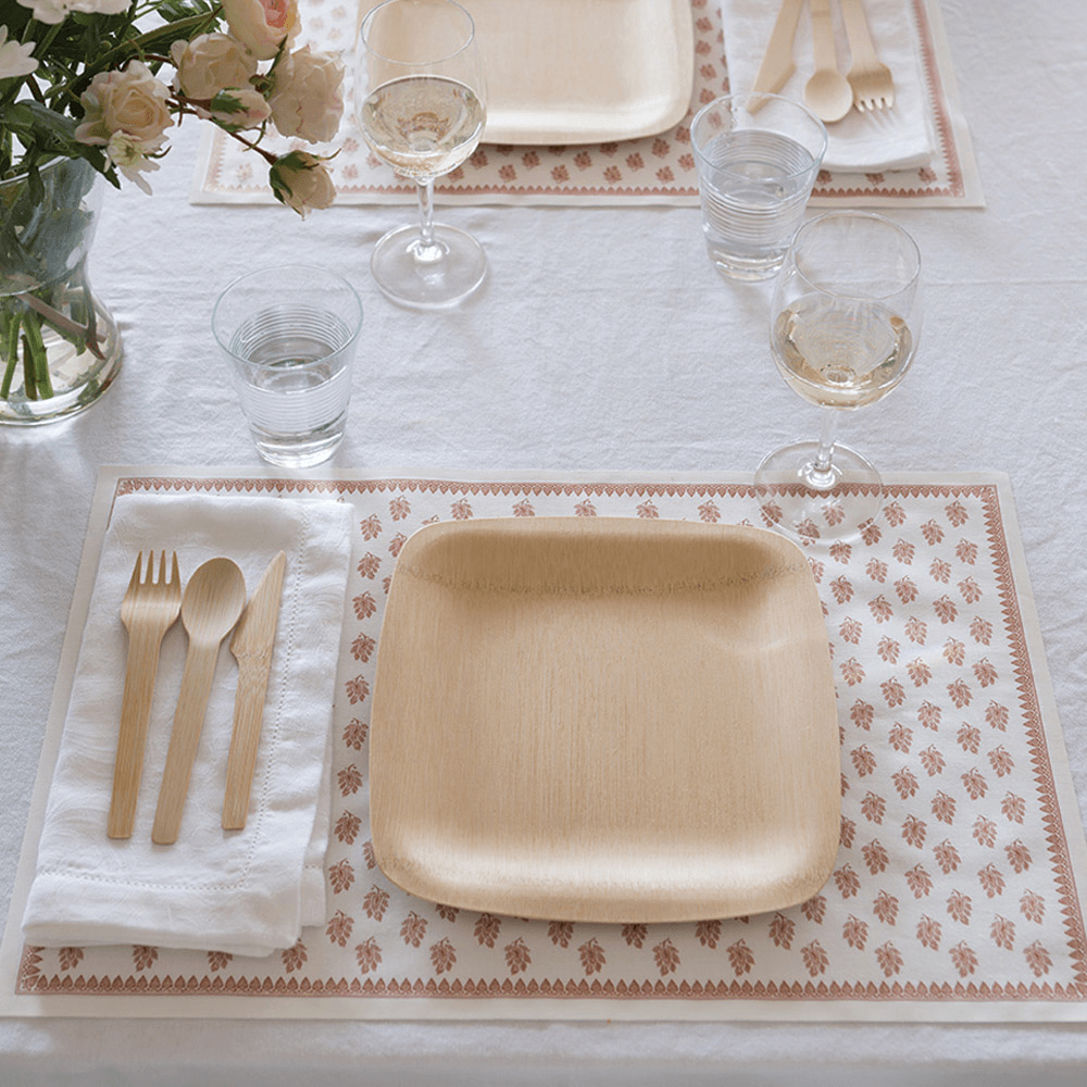 A place setting of a 9" square Veneerware plate, white cloth napkin, and Veneerware Cutlery are placed on a brown patterned placemat. 